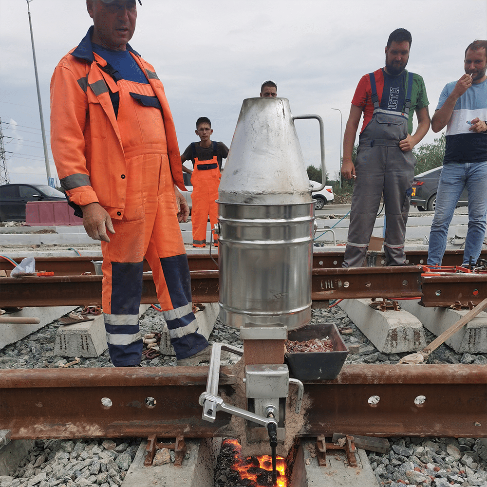 Aluminothermic rail welding casting stage: thermit crucible mounted above rail joint, molten steel pouring into mould, railway construction site