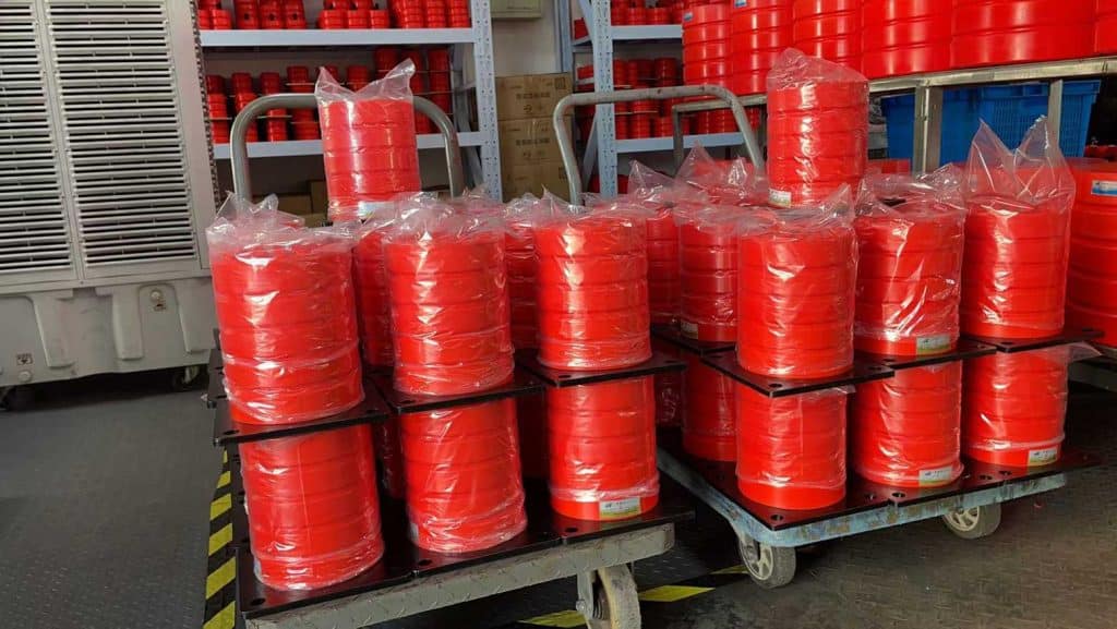 Warehouse stock photo of new red multi-layer polyurethane crane buffer bumpers with steel base plates, individually wrapped in plastic bags, stacked on metal trolleys; racks full of overhead crane rubber safety buffers in factory storage workshop, industrial lifting machinery spare parts