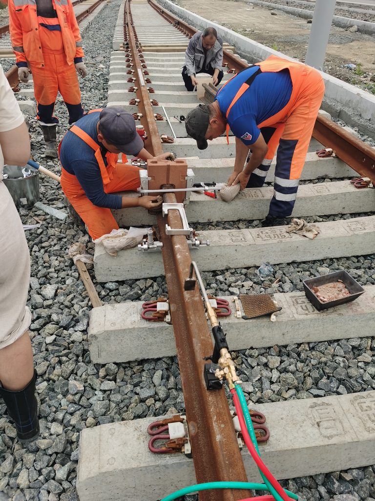 Workers installing mould for aluminothermic rail welding on railway track, preparing rail joint before preheating. Construction site with concrete sleepers and ballast.